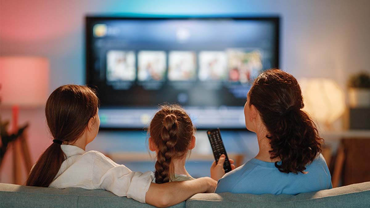 Mãe e duas filhas a ver televisão sentadas no sofá; vista de trás